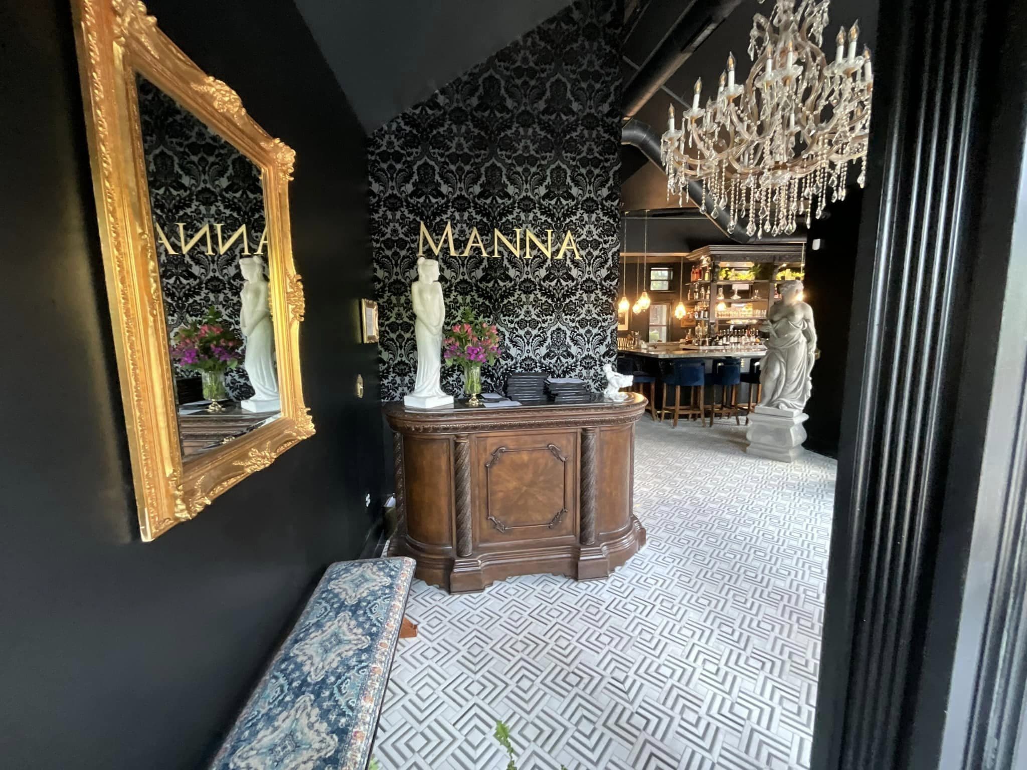 Remodeled entry at Manna in Columbus: black damask feature wall with gold lettering, a carved host stand, patterned tile floor and a crystal chandelier over the bar beyond.